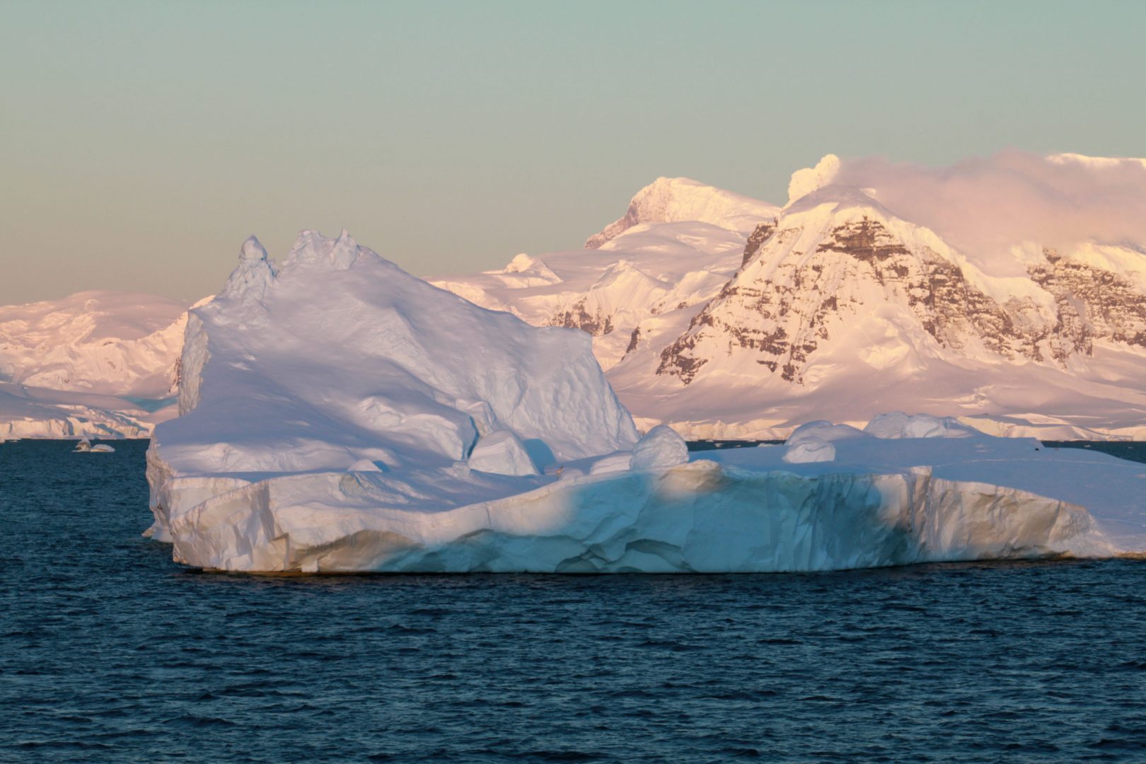 Zodiac boat cruising past icebergs with snow-covered Antarctic peaks in the background, under a clear sky.